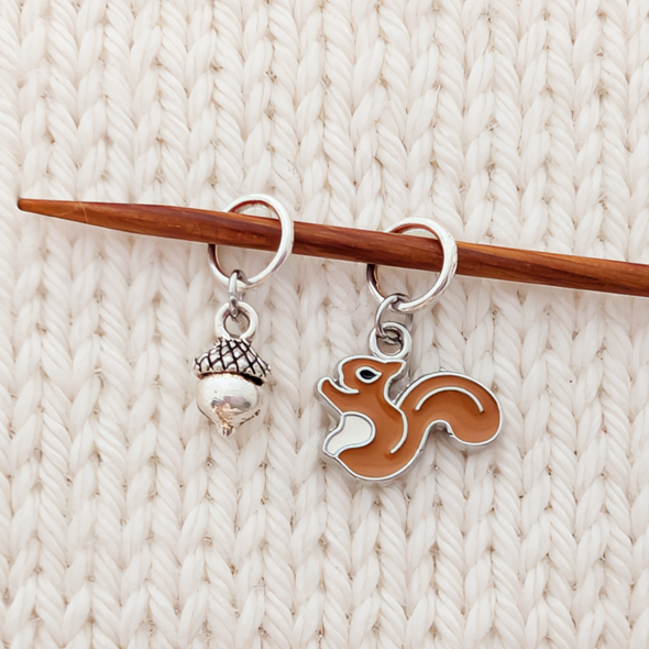 A close-up of the enamel squirrel and silver acorn ring stitch markers hanging from a wooden knitting needle, resting on cream colored knitting