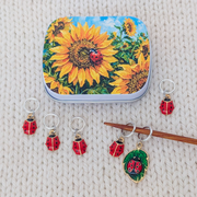 A notions tin and set of 6 ladybug-themed knitting stitch markers displayed on a cream knit blanket. The tin lid features a vibrant sunflower and ladybug illustration.