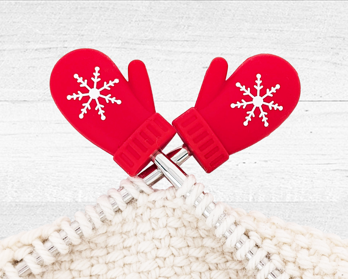 Pretty Warm Designs' red Christmas Mitten Stitch Stoppers shown on knitting needles, preventing the stitches of a chunky white yarn project from falling off.