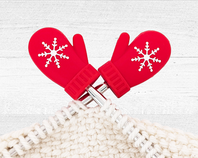 Pretty Warm Designs' red Christmas Mitten Stitch Stoppers shown on knitting needles, preventing the stitches of a chunky white yarn project from falling off.