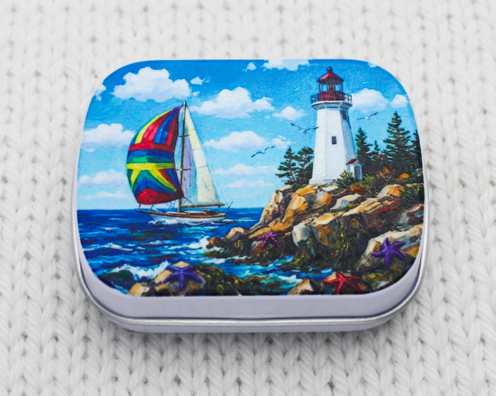 Notions storage tin for knitting featuring a coastal lighthouse and sailboat, resting on white knitted fabric.