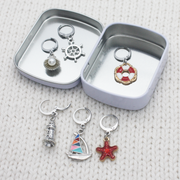 Travel-friendly hinged storage tin for fiber arts accessories, displaying a complete six-piece set of coastal theme clasp markers.