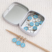 Set of ten blue on gold-toned number charm ring stitch markers on bamboo knitting needle with tin displayed on white knitted square by Pretty Warm Designs