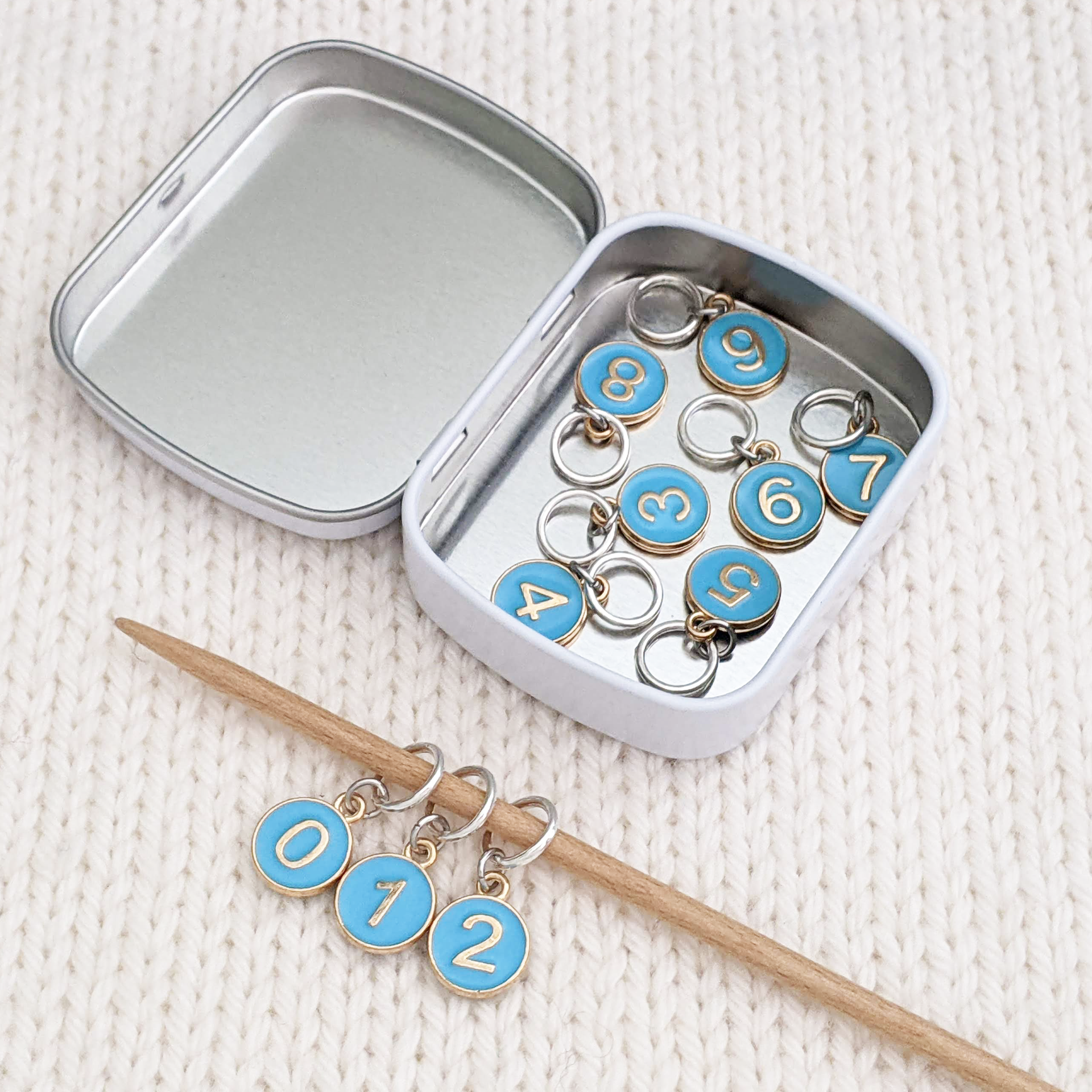 Set of ten blue on gold-toned number charm ring stitch markers on bamboo knitting needle with tin displayed on white knitted square by Pretty Warm Designs