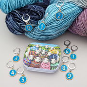 A set of locking stitch markers with gold-tone edges and blue centers numbered 0 through 9, shown with silver-tone clips. The markers are styled on variegated yarn in blue, teal, and purple, with a small illustrated tin box in the foreground, the sheep sweater club artwork.