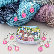 A set of locking stitch markers and a small storage tin arranged on a light surface with a skein of yarn in the background. The markers are round pink discs with gold numbers 0 through 9, attached to silver stitch holders.