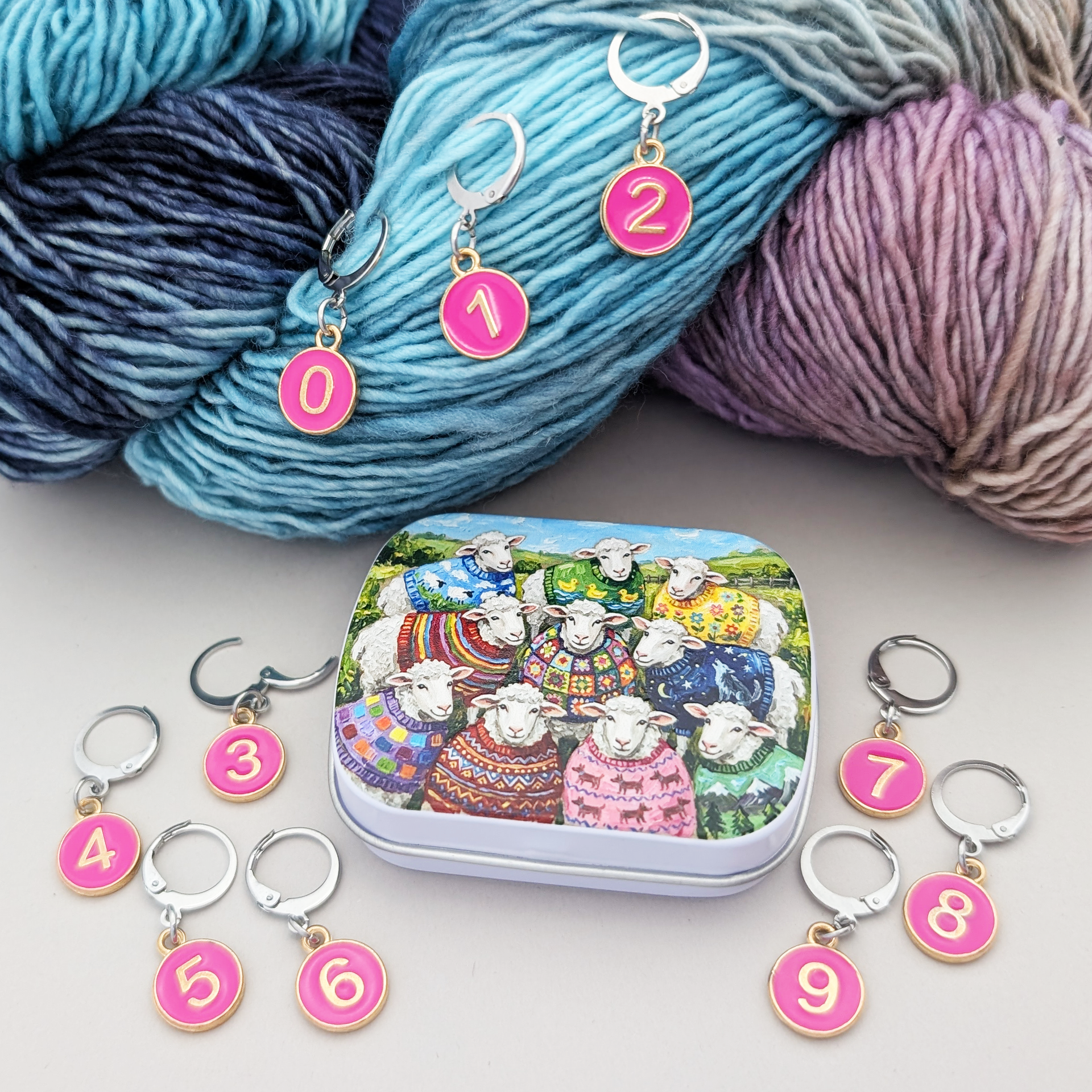 A set of locking stitch markers and a small storage tin arranged on a light surface with a skein of yarn in the background. The markers are round pink discs with gold numbers 0 through 9, attached to silver stitch holders.