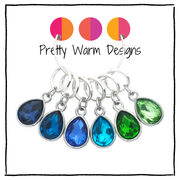 Six silver ring charms with teardrop-shaped faceted gemstones in shades of blue and green, with the brand name 'Pretty Warm Designs' 