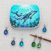 A fiber arts notions tin with an humpback whale and calf print and a set of 6 ring stitch makers for knitting with teardrop crystal glass charms in blues and greens