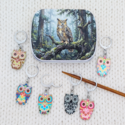 A set of owl-themed removable stitch holders arranged around a small hinged tin with artwork depicting an owl in a forest. The progress keepers feature colorful owl faces and bodies in pink, blue, yellow, black, and white, with silver rings.