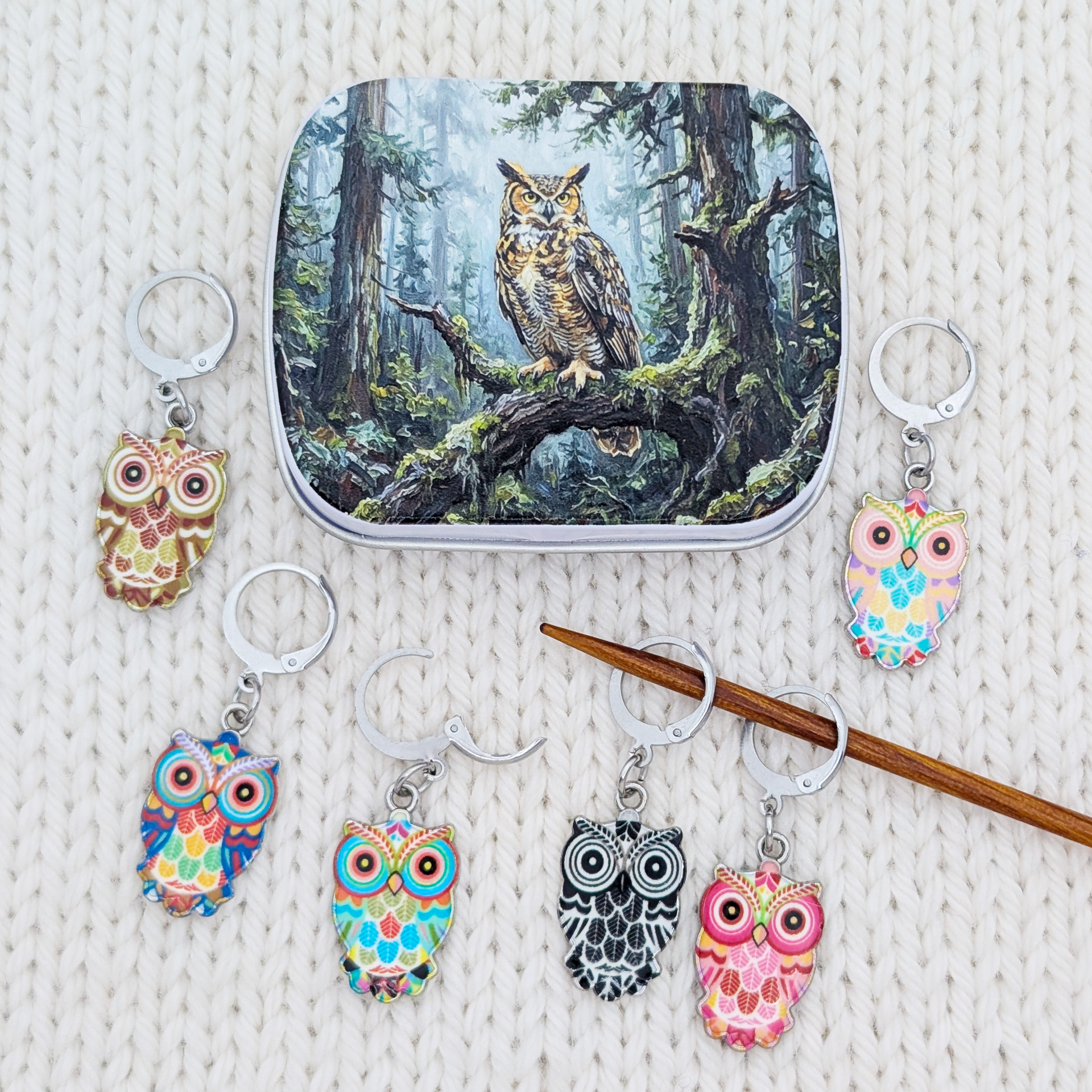 A set of owl-themed removable stitch holders arranged around a small hinged tin with artwork depicting an owl in a forest. The progress keepers feature colorful owl faces and bodies in pink, blue, yellow, black, and white, with silver rings.
