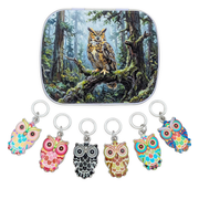 A square metal tin lid featuring an illustration of a large owl perched on a mossy tree branch in a forest, shown above six owl-shaped ring style stitch markers in different colorful patterns on silver rings.