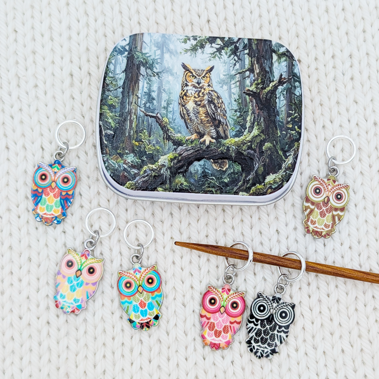 A set of owl-themed knitting stitch markers and a matching metal tin featuring an owl in a forest. The stitch markers come in various colorful and black-and-white owl patterns, arranged on a white textured knit blanket.