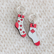 Pair of handmade wooden Canada Flag Socks knitting stitch markers with snag-free closed silver rings on a cream knit background. One sock charm features a bold red maple leaf, and the other shows a scattered maple leaf pattern.