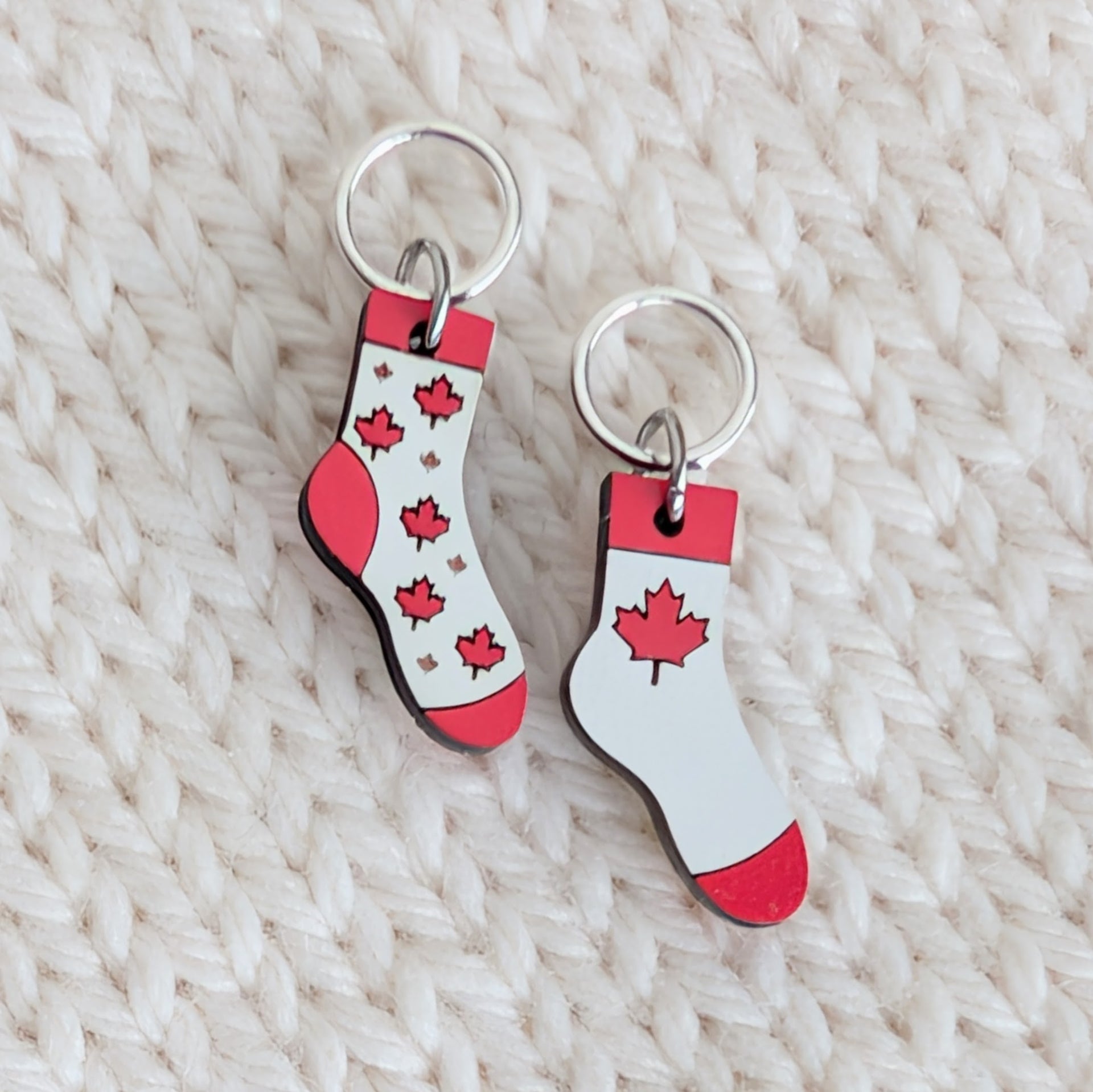 Pair of handmade wooden Canada Flag Socks knitting stitch markers with snag-free closed silver rings on a cream knit background. One sock charm features a bold red maple leaf, and the other shows a scattered maple leaf pattern.