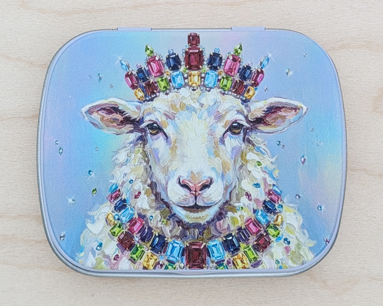 A hinged metal notions tin featuring a painted illustration of a white sheep wearing a colorful jeweled crown and necklace, set against a light blue background, photographed on a light wood surface.