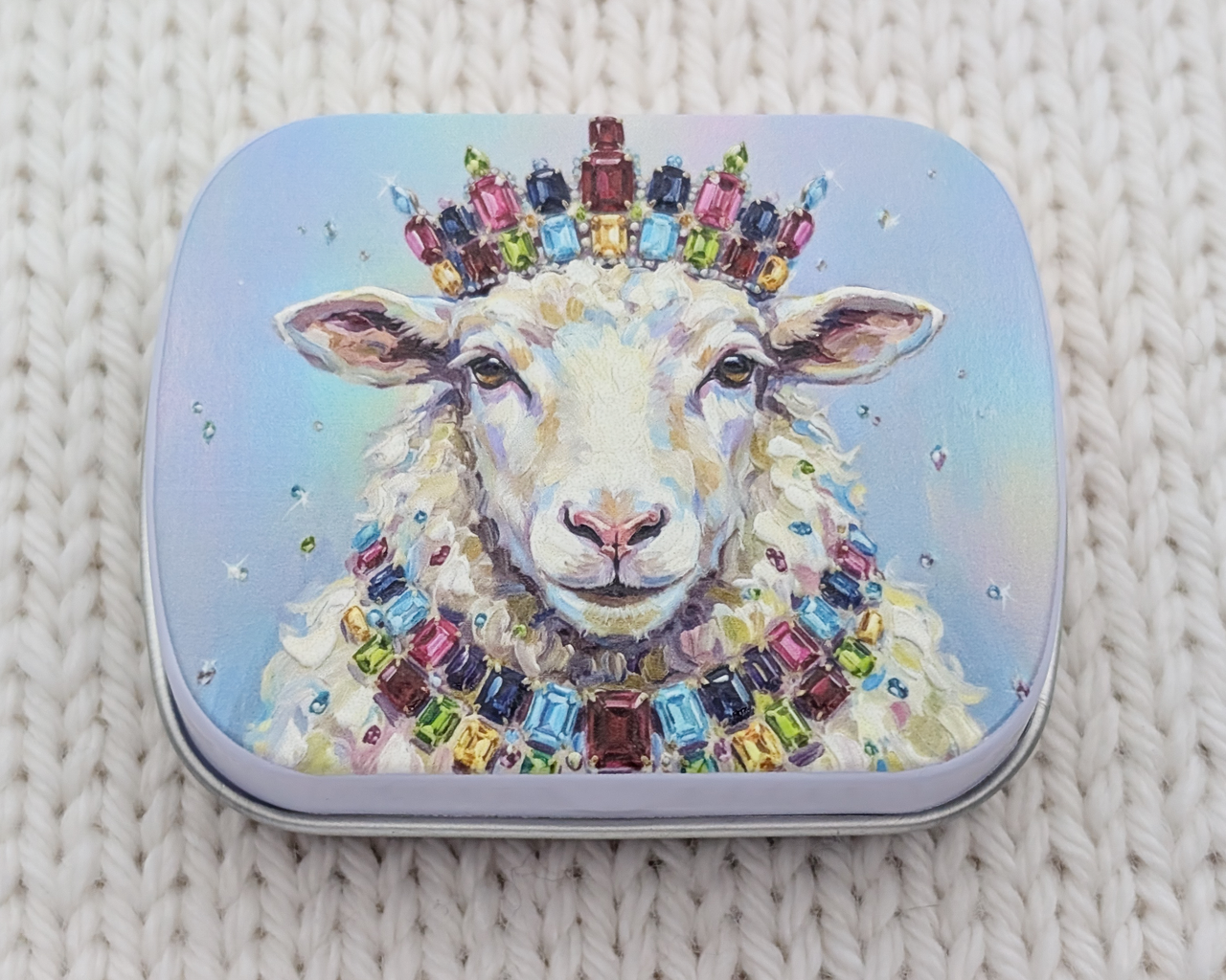 A hinged metal notions tin featuring a colorful illustration of a white sheep wearing a jeweled crown and necklace, placed on a textured cream knit blanket.
