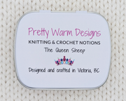 The bottom side of a hinged metal notions tin with printed text that reads 'Pretty Warm Designs KNITTING & CROCHET NOTIONS, The Queen Sheep' and 'Designed and crafted in Victoria, BC' with a small jeweled crown graphic. The tin is placed on a cream-colored chunky knit fabric background.