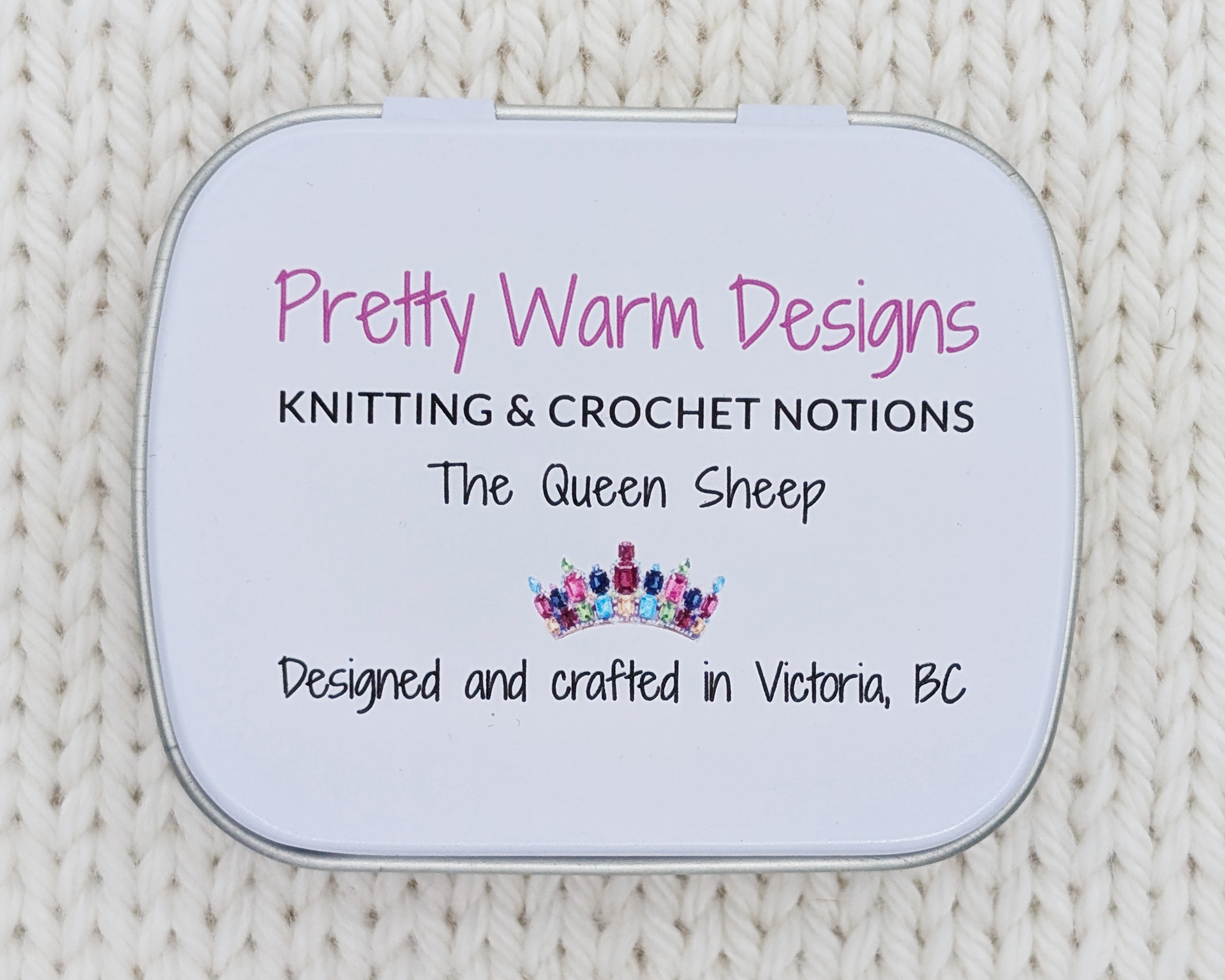 The bottom side of a hinged metal notions tin with printed text that reads 'Pretty Warm Designs KNITTING & CROCHET NOTIONS, The Queen Sheep' and 'Designed and crafted in Victoria, BC' with a small jeweled crown graphic. The tin is placed on a cream-colored chunky knit fabric background.