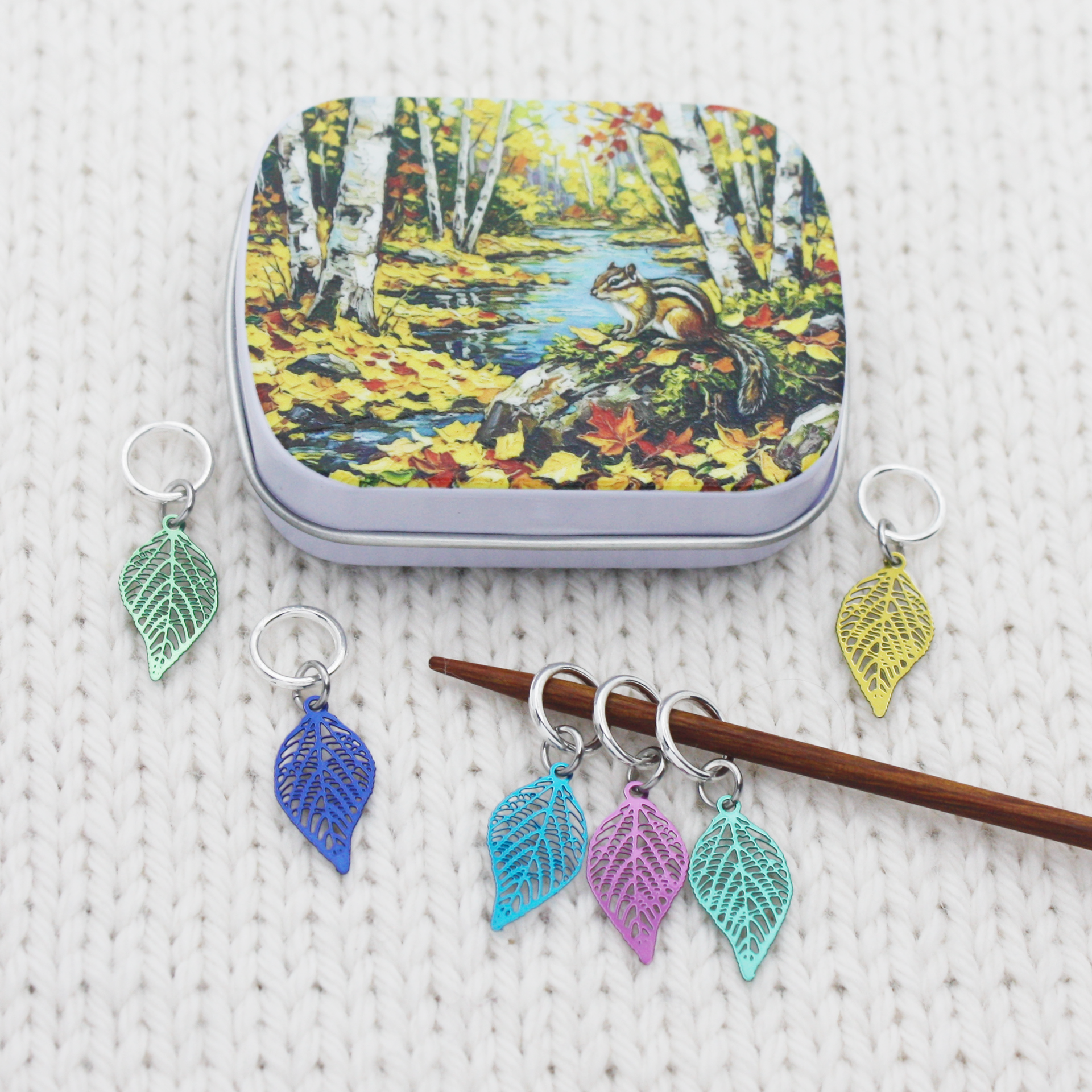 A small craft storage notions tin with a chipmunk in an autumn scene with six ring style stitch markers for knitters, snag-free closed rings with lightweight filigree metal charms