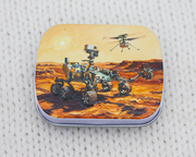 A notions tin with artwork of NASA's Perseverance rover on Mars, a dusty orange desert landscape under a pale yellow sky, resting on a white textured knit surface.