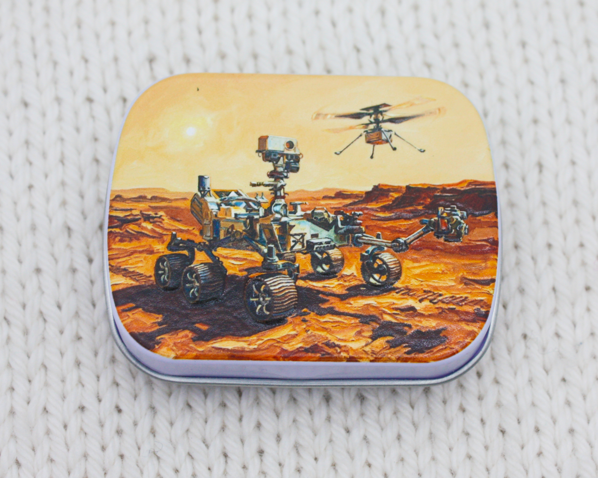 A notions tin with artwork of NASA's Perseverance rover on Mars, a dusty orange desert landscape under a pale yellow sky, resting on a white textured knit surface.