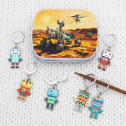 A set of colorful robot locking stitch markers arranged around a small notions tin featuring a Mars rover illustration. 