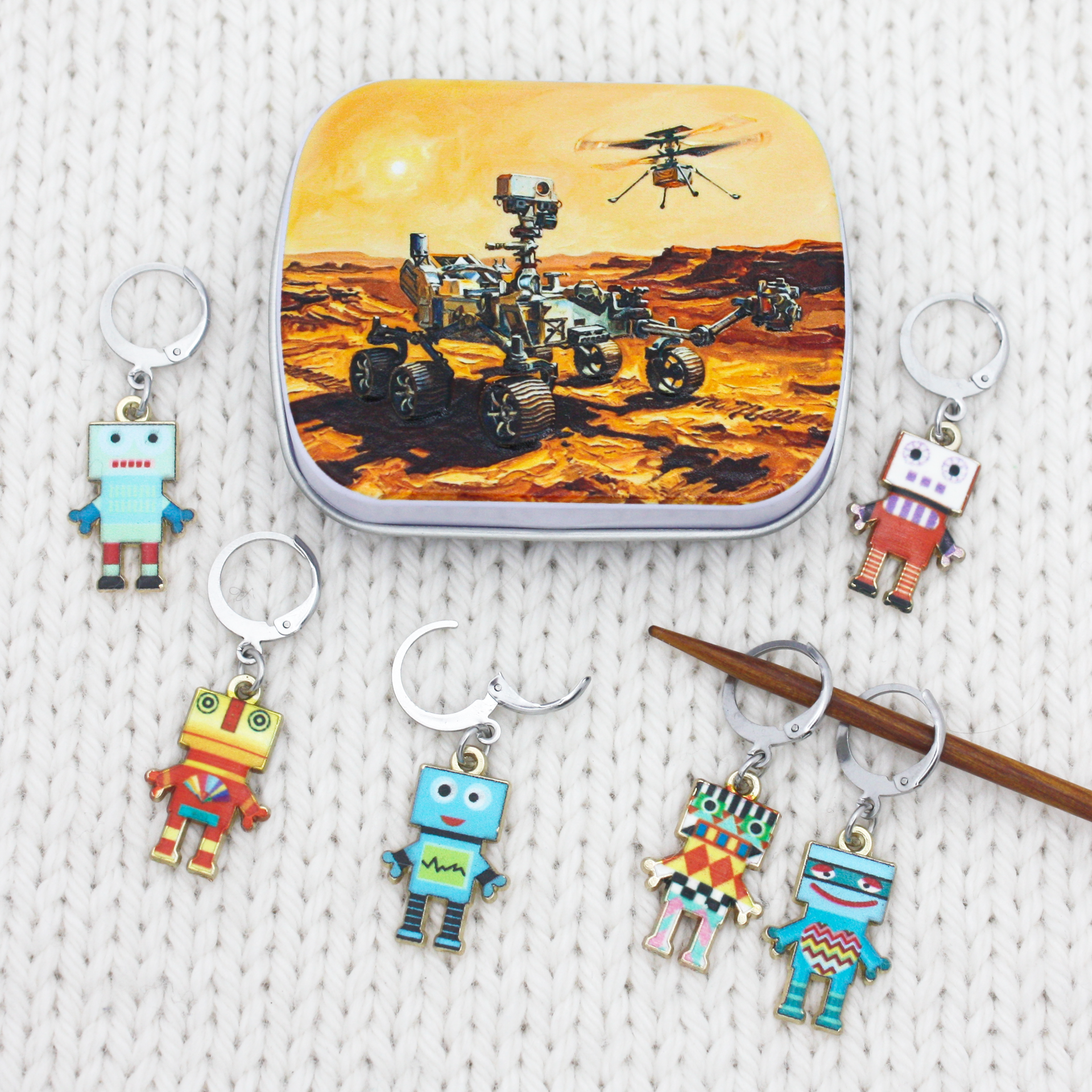 A set of colorful robot locking stitch markers arranged around a small notions tin featuring a Mars rover illustration. 