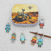 A set of colorful robot-shaped ring stitch markers arranged on a white knit fabric background, with a small notions tin featuring a rover illustration at the top. The charms have silver metal rings and are shown in multiple bright color combinations.