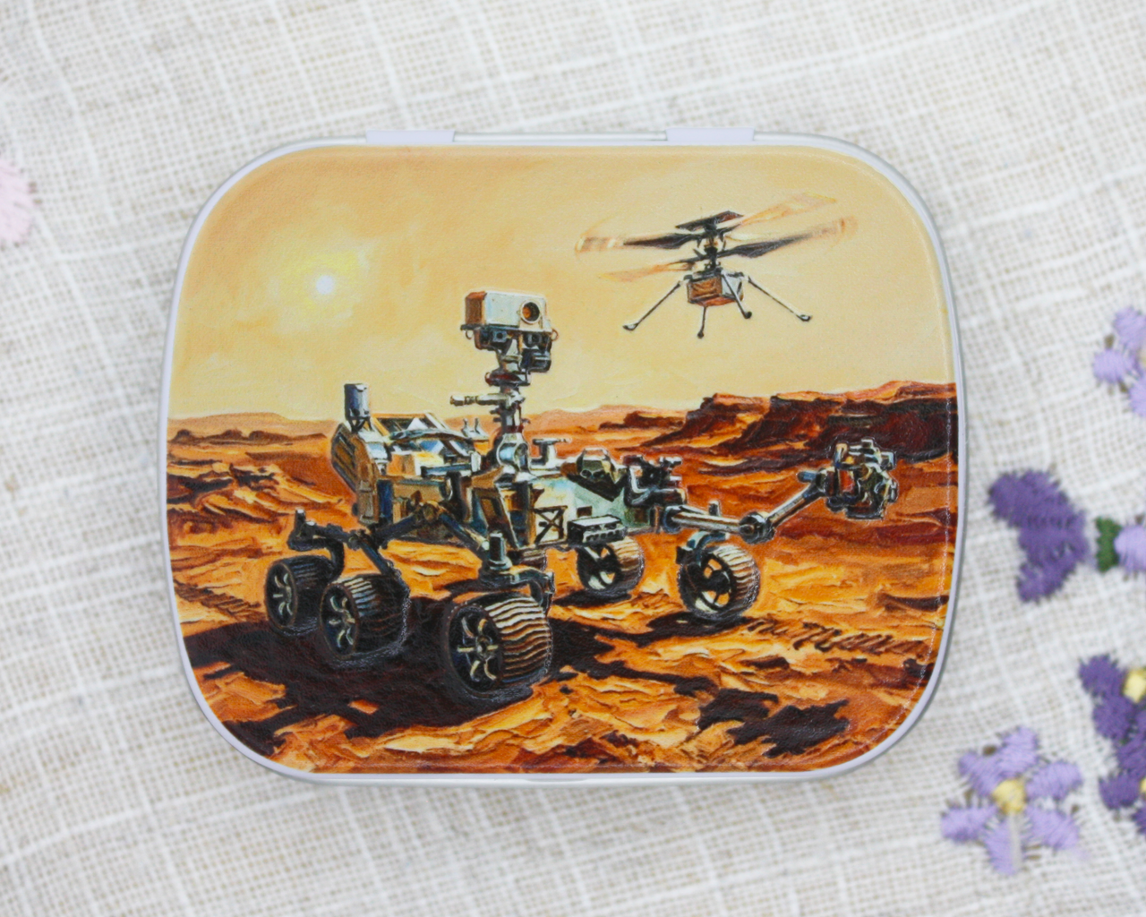 A notions tin with artwork of NASA's Perseverance rover on Mars, a dusty orange desert landscape under a pale yellow sky.
