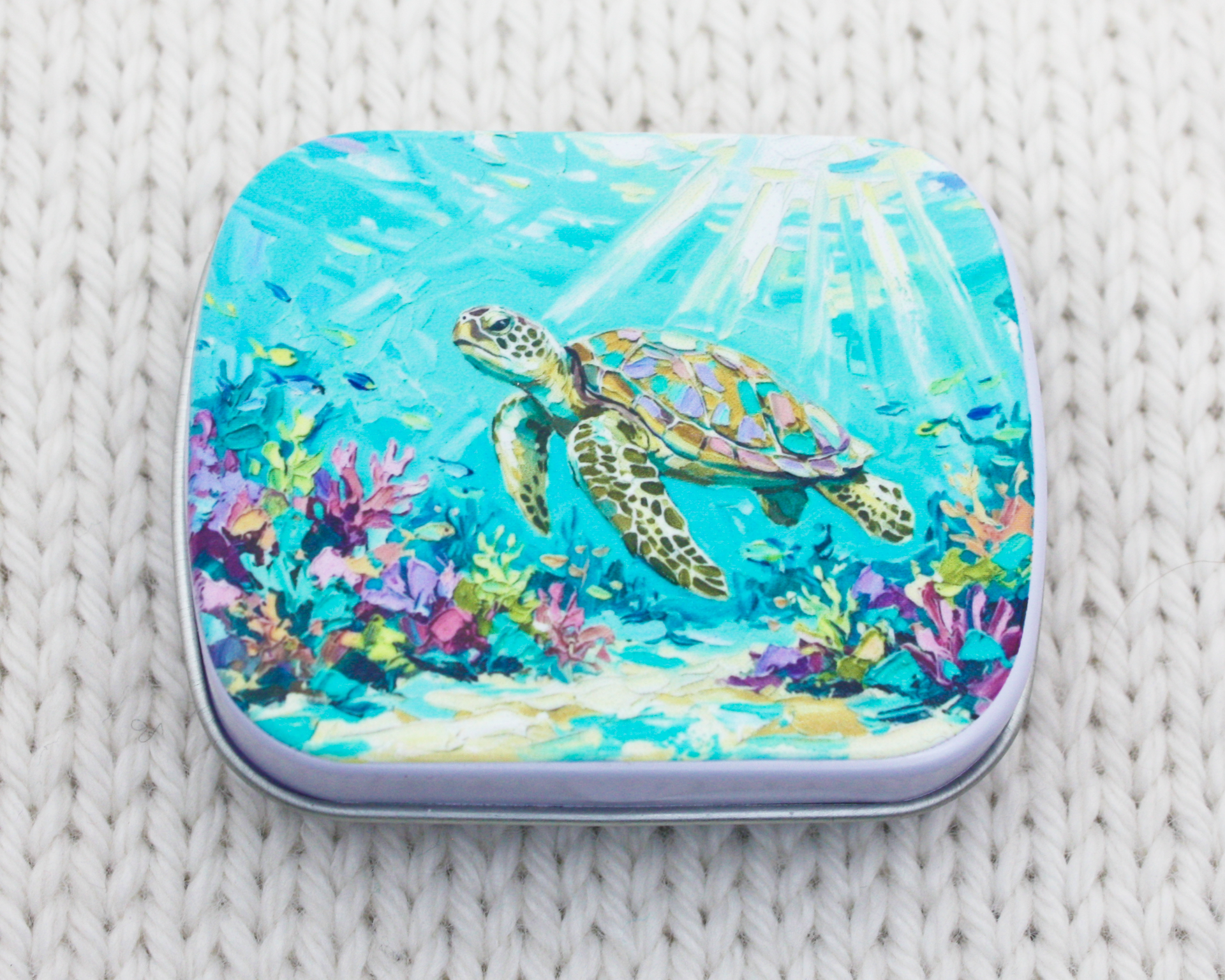 A metal notions case for stitch markers featuring an underwater illustration of a sea turtle swimming over coral reefs