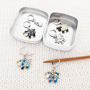 Sheep locking stitch markers styled in and around an open Pretty Warm Designs tin on a cream knitted background, with a bamboo needle, featuring a Valais Blacknose, a silver lamb,  two colourful patchwork sheep, an I love yarn charm and a black and white curly sheep.