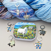 A set of sheep-themed crochet and knitting stitch markers with various colorful and silver designs, arranged around a small tin featuring a sheep in a field, with blue variegated yarn in the background.