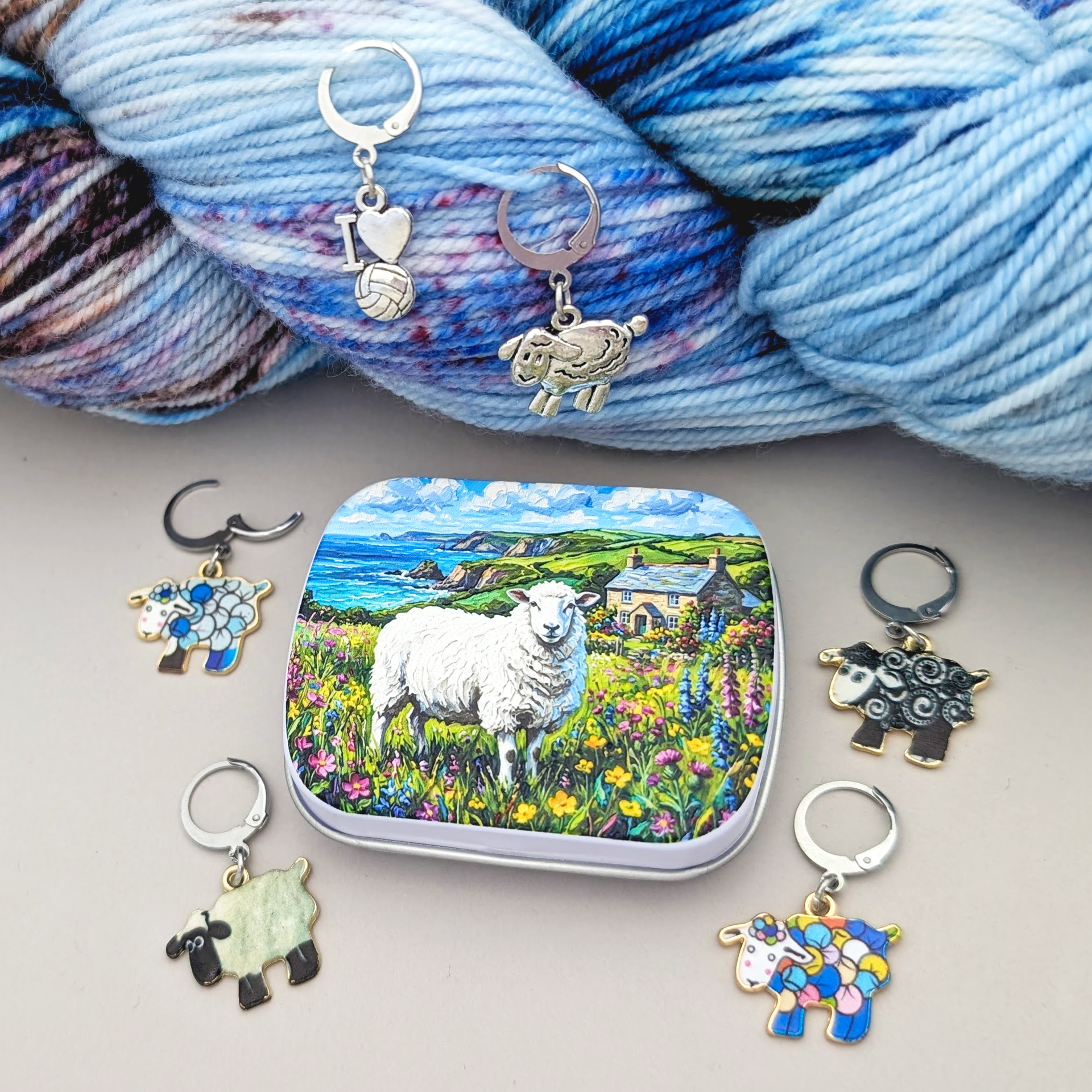 A set of sheep-themed crochet and knitting stitch markers with various colorful and silver designs, arranged around a small tin featuring a sheep in a field, with blue variegated yarn in the background.