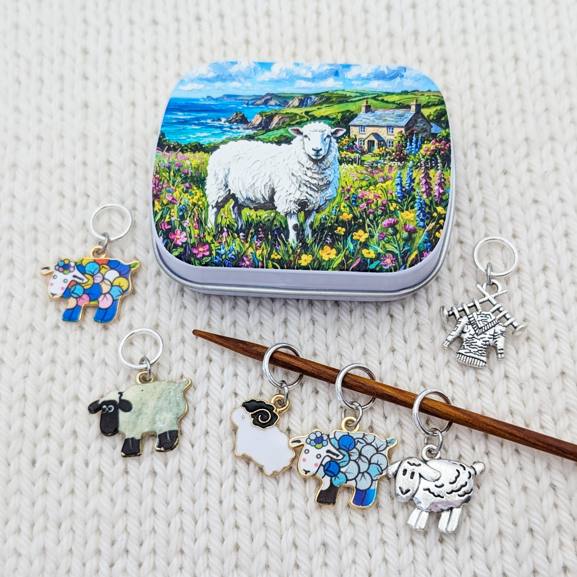 A set of sheep knitting stitch markers and a matching tin featuring a colorful illustration of a white sheep in a flower field with a blue sea and cottage background, arranged on a white knitted fabric.