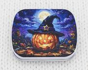 A small storage tin tfeaturing a Halloween illustration of a glowing jack-o'-lantern wearing a witch hat under a full moon
