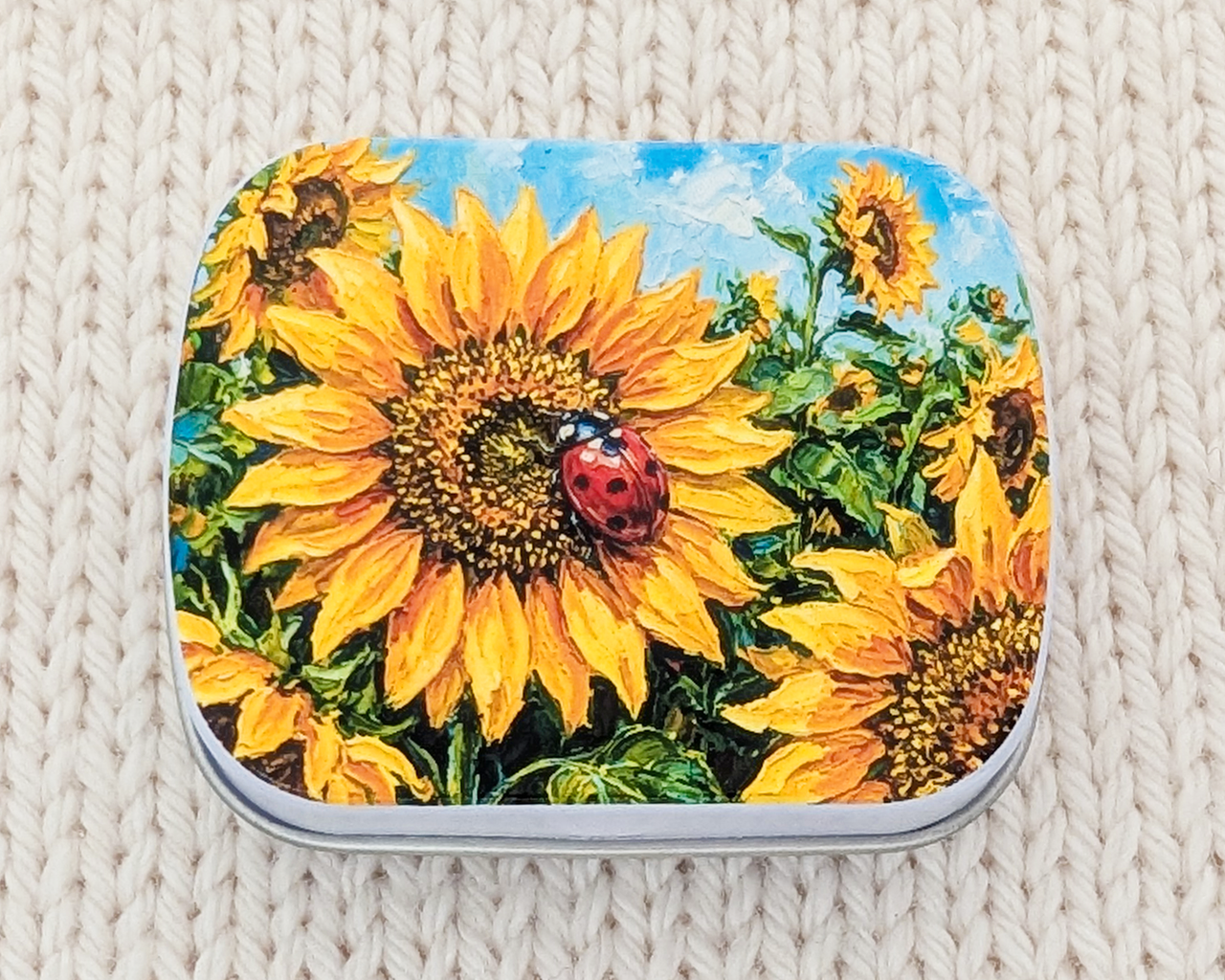 A metal notions tin for stitch marker storage with yellow sunflowers and a red ladybug on the lid
