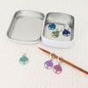 Six colour coated stainless steel round charm knitting stitch markers on knitting needle sitting on white yarn knitted background with storage tin by Pretty Warm Designs Inc