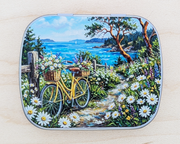 A hinged metal notions tin featuring a colorful scenic illustration of a yellow bicycle on a flower-lined path near the ocean, photographed on a light wood surface.