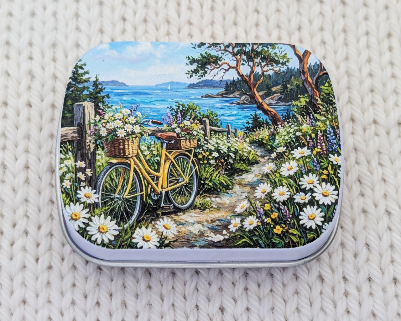A hinged metal notions tin featuring a colorful landscape illustration of a yellow bicycle on a flower-lined path near the ocean, placed on a cream-colored knitted blanket.