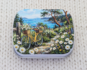 A hinged metal notions tin featuring a colorful landscape illustration of a yellow bicycle on a flower-lined path near the ocean, placed on a cream-colored knitted blanket.
