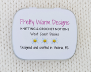 The bottom of a hinged metal notions tin  printed with the brand name 'Pretty Warm Designs' and 'Knitting & Crochet Notions', along with 'West Coast Daisies' and small daisy illustrations, Designed and crafted in Victoria BC, placed on a cream-colored knitted fabric background.