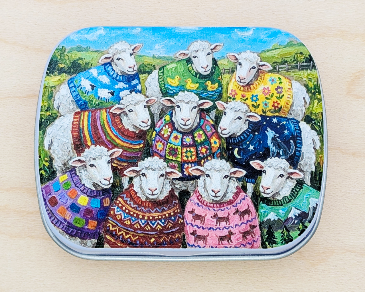 A hinged metal notions tin featuring a colorful illustration of ten sheep wearing vibrant, patterned knit and crochet sweaters in a grassy field under a blue sky. The tin is placed on a light wood surface.