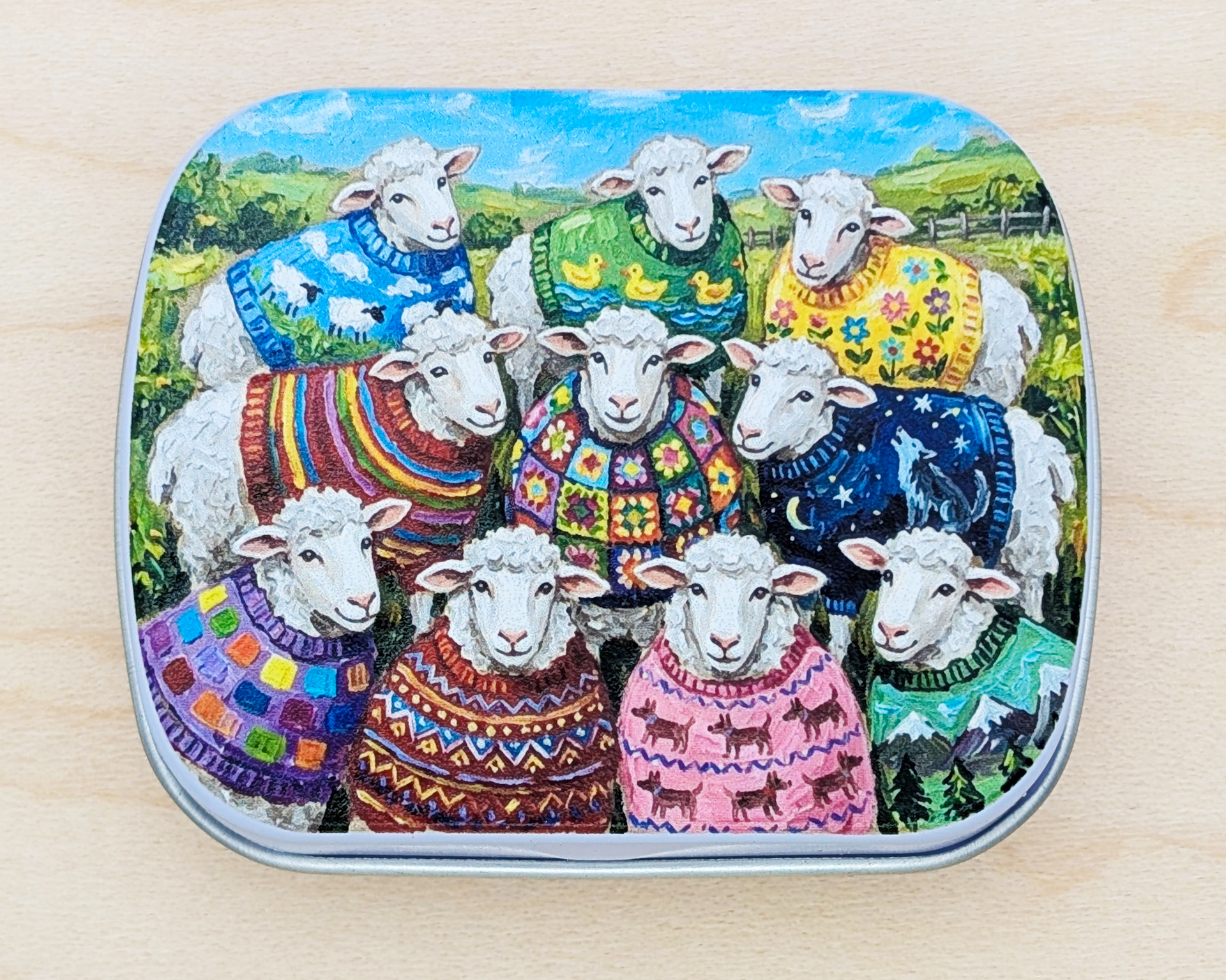 A hinged metal notions tin featuring a colorful illustration of ten sheep wearing vibrant, patterned knit and crochet sweaters in a grassy field under a blue sky. The tin is placed on a light wood surface.
