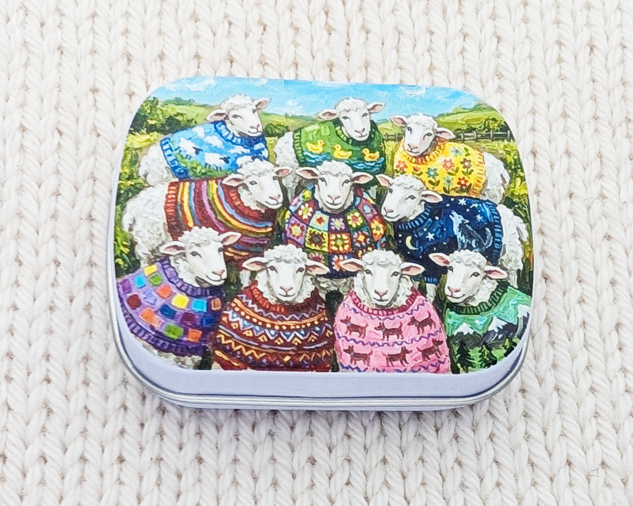 A hinged metal notions tin featuring a colorful illustration of sheep wearing knitted sweaters in a landscape, placed on a cream-colored textured knit blanket.