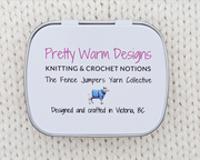 The bottom side of a hinged metal notions tin, placed on a cream-colored knitted blanket. The bottom features printed text reading 'Pretty Warm Designs' and 'Knitting & Crochet Notions' "designed and crafted in Victoria, BC' with a small illustration of a sheep.