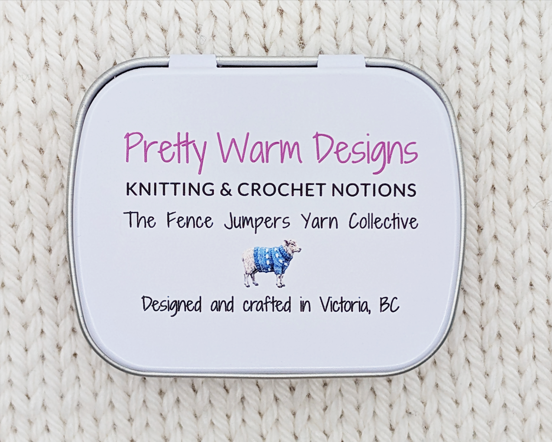 The bottom side of a hinged metal notions tin, placed on a cream-colored knitted blanket. The bottom features printed text reading 'Pretty Warm Designs' and 'Knitting & Crochet Notions' "designed and crafted in Victoria, BC' with a small illustration of a sheep.