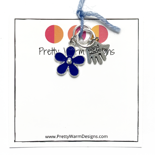 Two knitting stitch markers include one blue flower and one silver plated hand stamped with Hand Made charm attached with blue yarn to white cardstock printed with Pretty Warm Designs logo and text