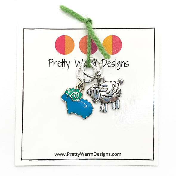 Two knitting ring stitch markers, one enamel blue and green sheep and one Tibetan antiqued silver lamb attached with green yarn to cardstock with Pretty Warm Designs text and logo