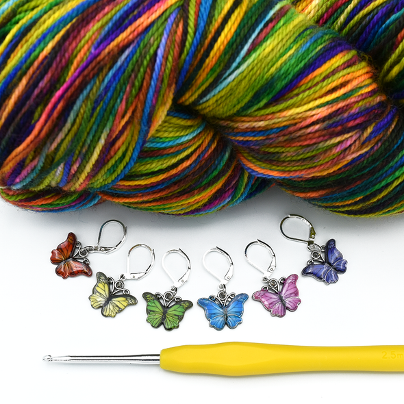 Set of six enamel butterfly charm crochet locking stitch markers in red, yellow, green, blue, pink and purple with brightly coloured variegated yarn and yellow handled crochet hook for crochet by Pretty Warm Designs